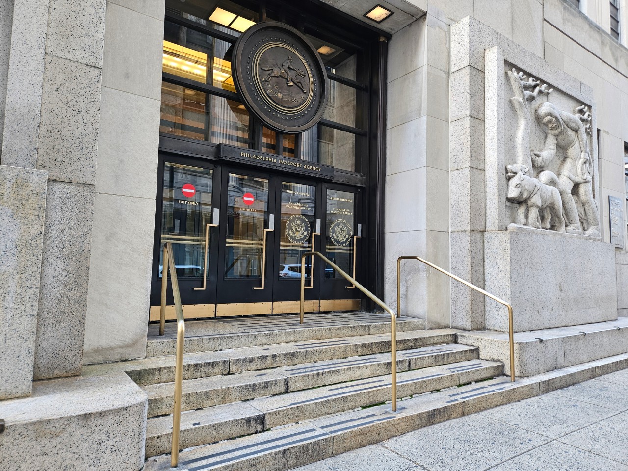 Exterior of Philadelphia Passport Agency building. It is a beige rectangular office building with artwork engraved near the entrance.