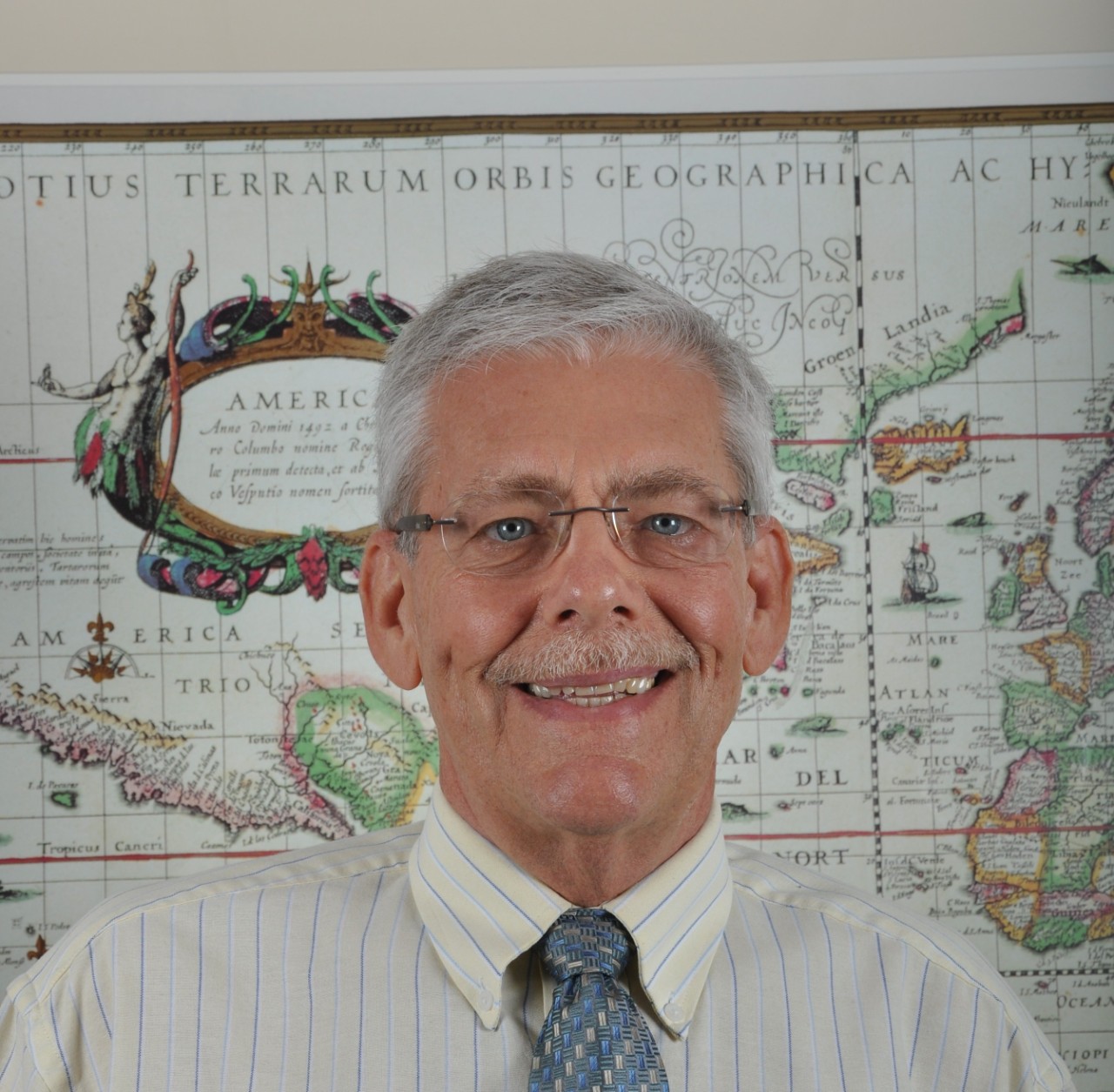 An elderly man standing in front of a map of the world hanging on a wall.